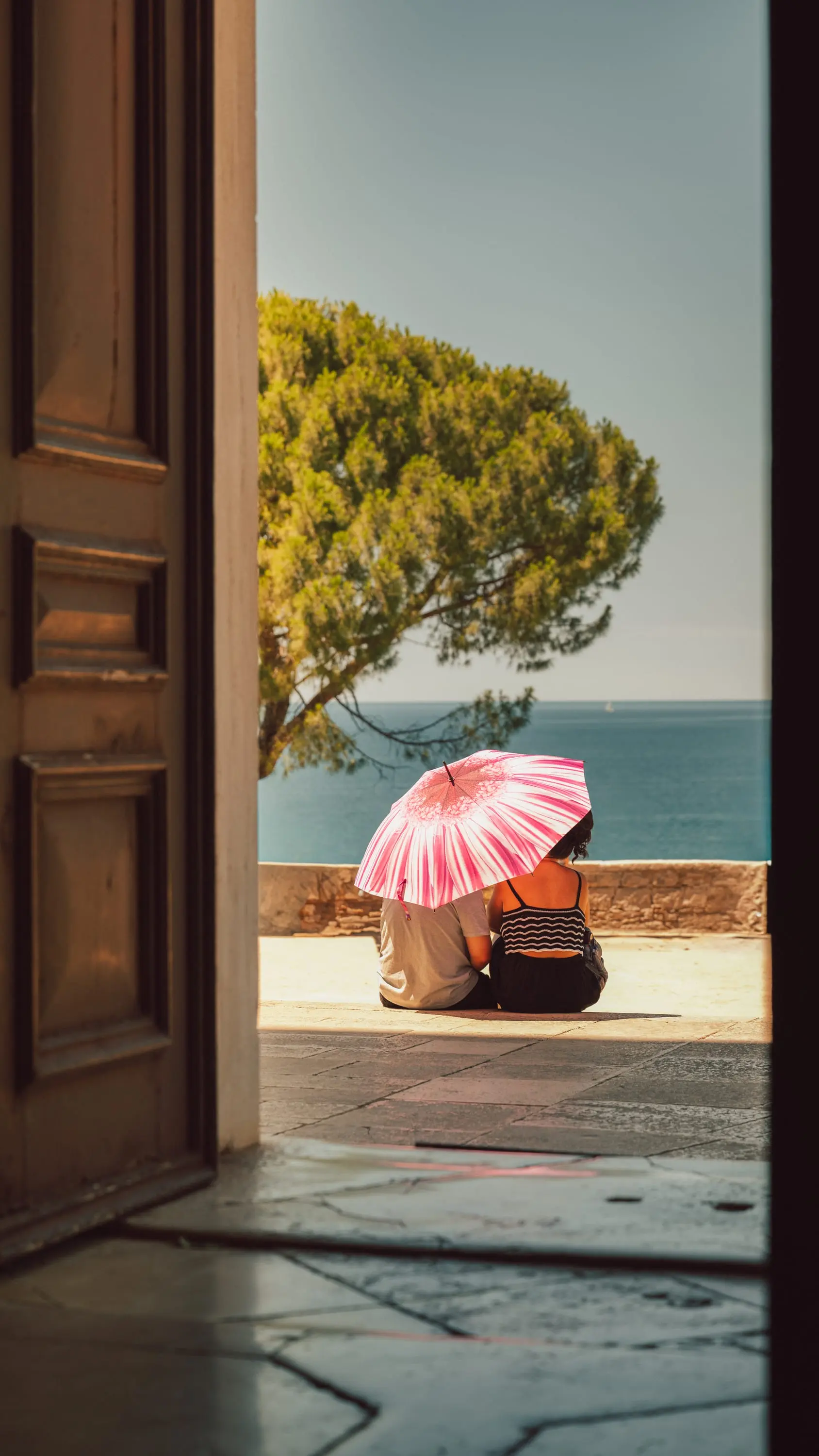 Couple under a pink parasol by the sea, framed by an archway